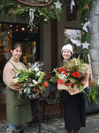 Two smiling Senka Florist team members holding festive winter hand-tied bouquets outside the flower shop decorated with holiday garlands in Whistler