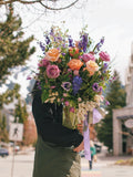 A florist holding a massive, vibrant floral arrangement with peach and magenta roses, purple wildflowers, and a lilac ribbon in Whistler village.