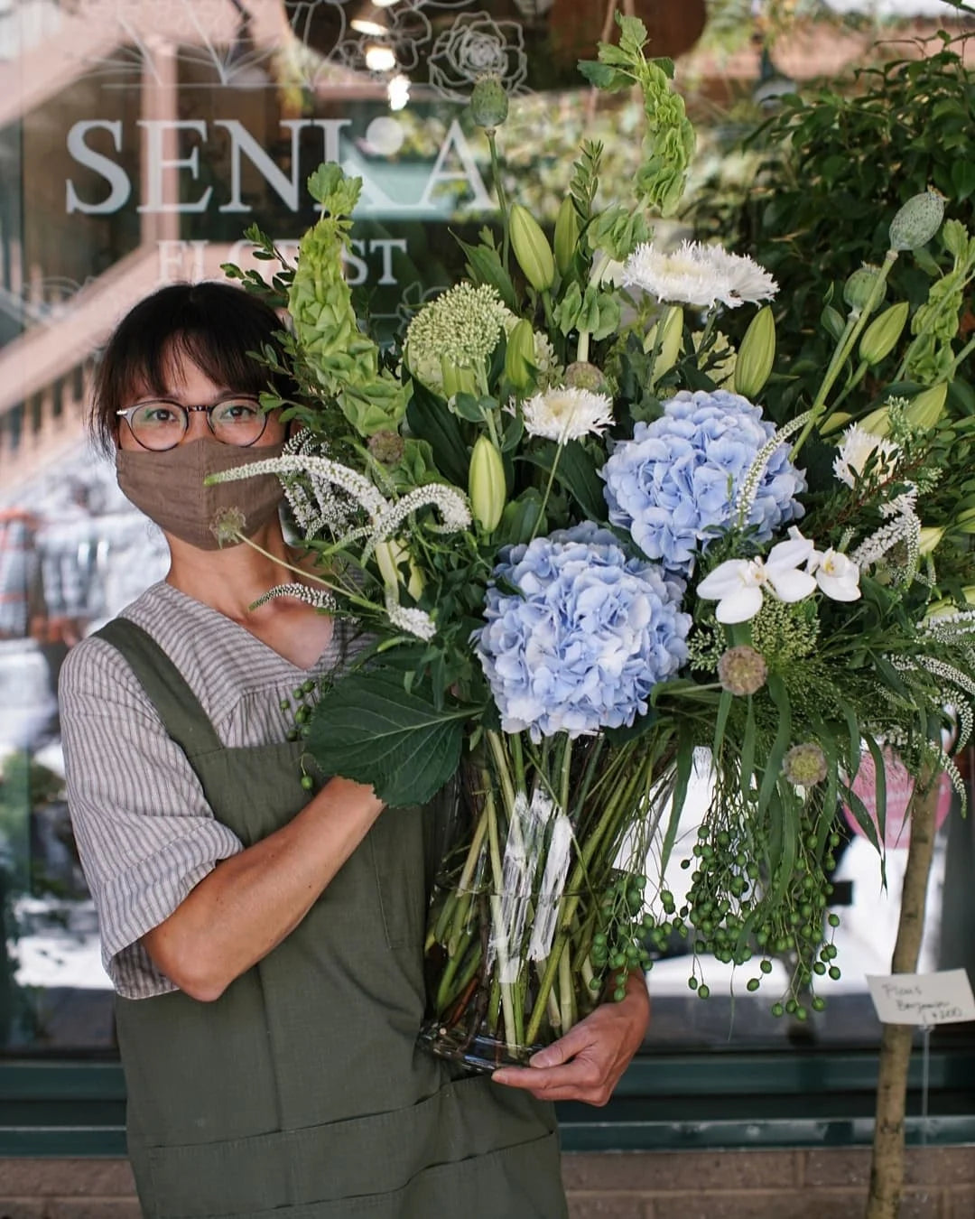 Person holding a large bouquet of flowers in front of a store named 'Senka'.