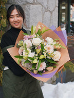 Smiling florist holding a large hand-tied bouquet with white roses, pink gerbera daisies, and purple accent flowers wrapped in brown and purple paper.
