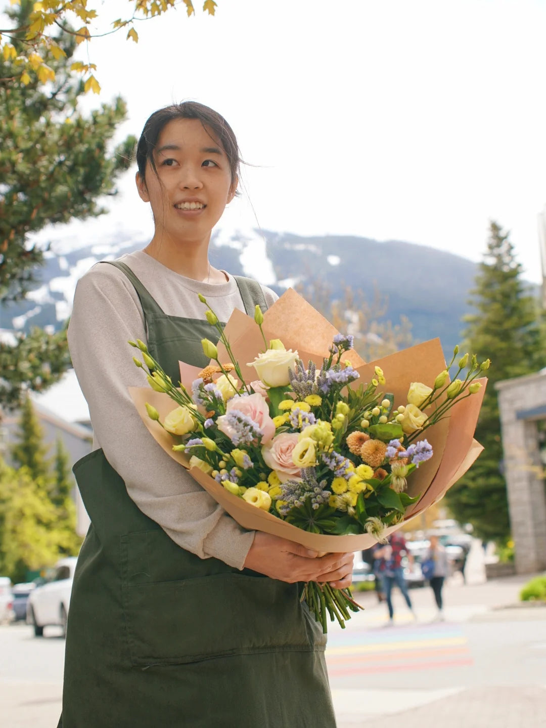 Senka Florist team member holding a large, vibrant yellow and pastel hand-tied bouquet with the Whistler mountains in the background.