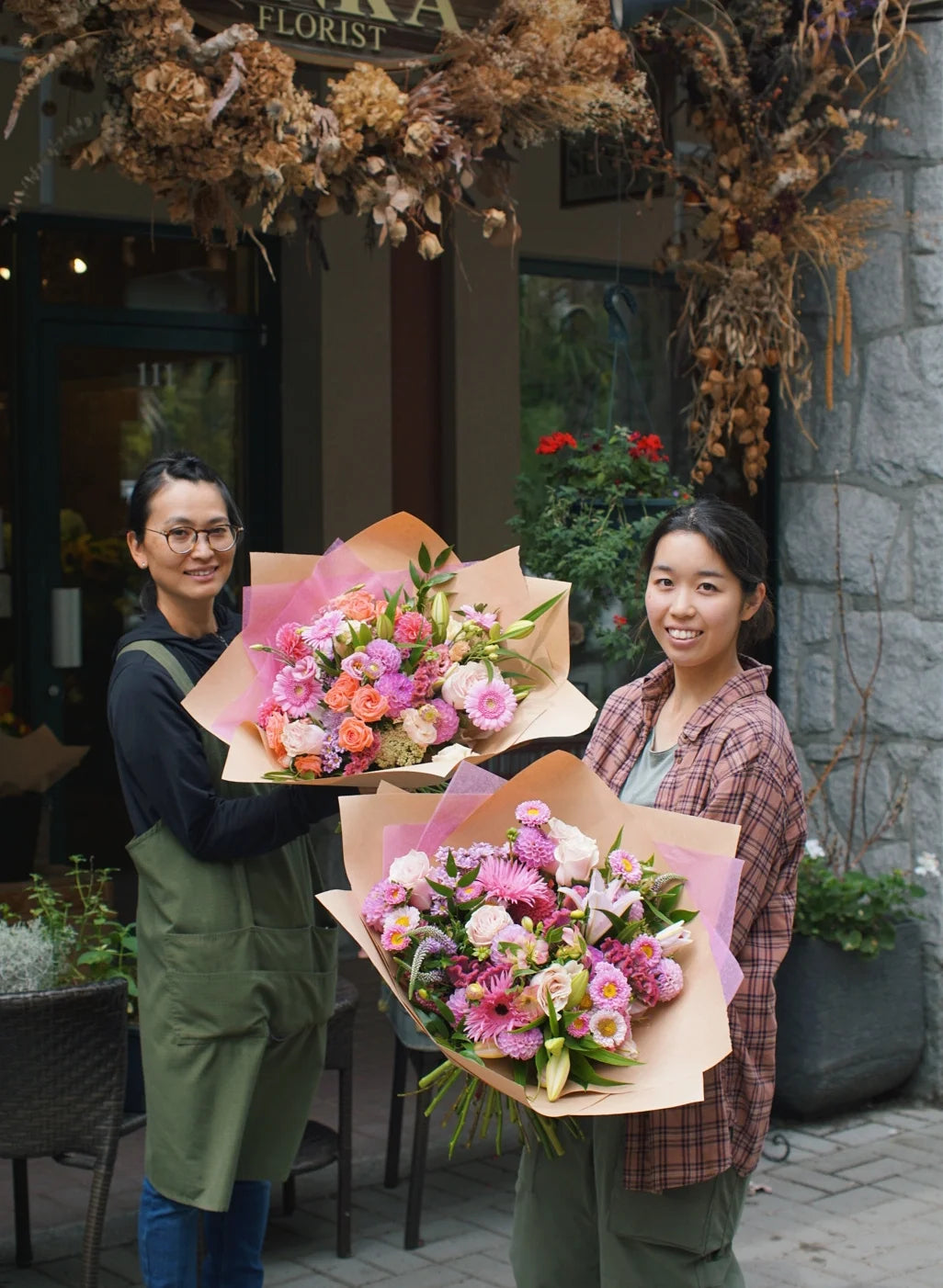 Two smiling Senka Florist team members holding large, vibrant pink hand-tied bouquets outside the flower shop in Whistler