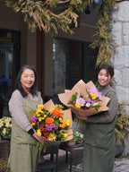 Two smiling florists holding a vibrant orange and yellow bouquet and a soft pastel flower arrangement outside the shop.