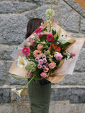 A vibrant, seasonal hand-tied flower arrangement with bright pink, coral, and white blooms wrapped in signature brown paper.