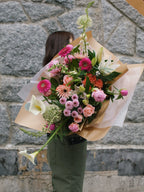 A vibrant, seasonal hand-tied flower arrangement with bright pink, coral, and white blooms wrapped in signature brown paper.