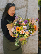 Smiling florist holding a vibrant hand-tied bouquet featuring orange gerbera daisies, peach roses, and purple accent flowers wrapped in brown paper.